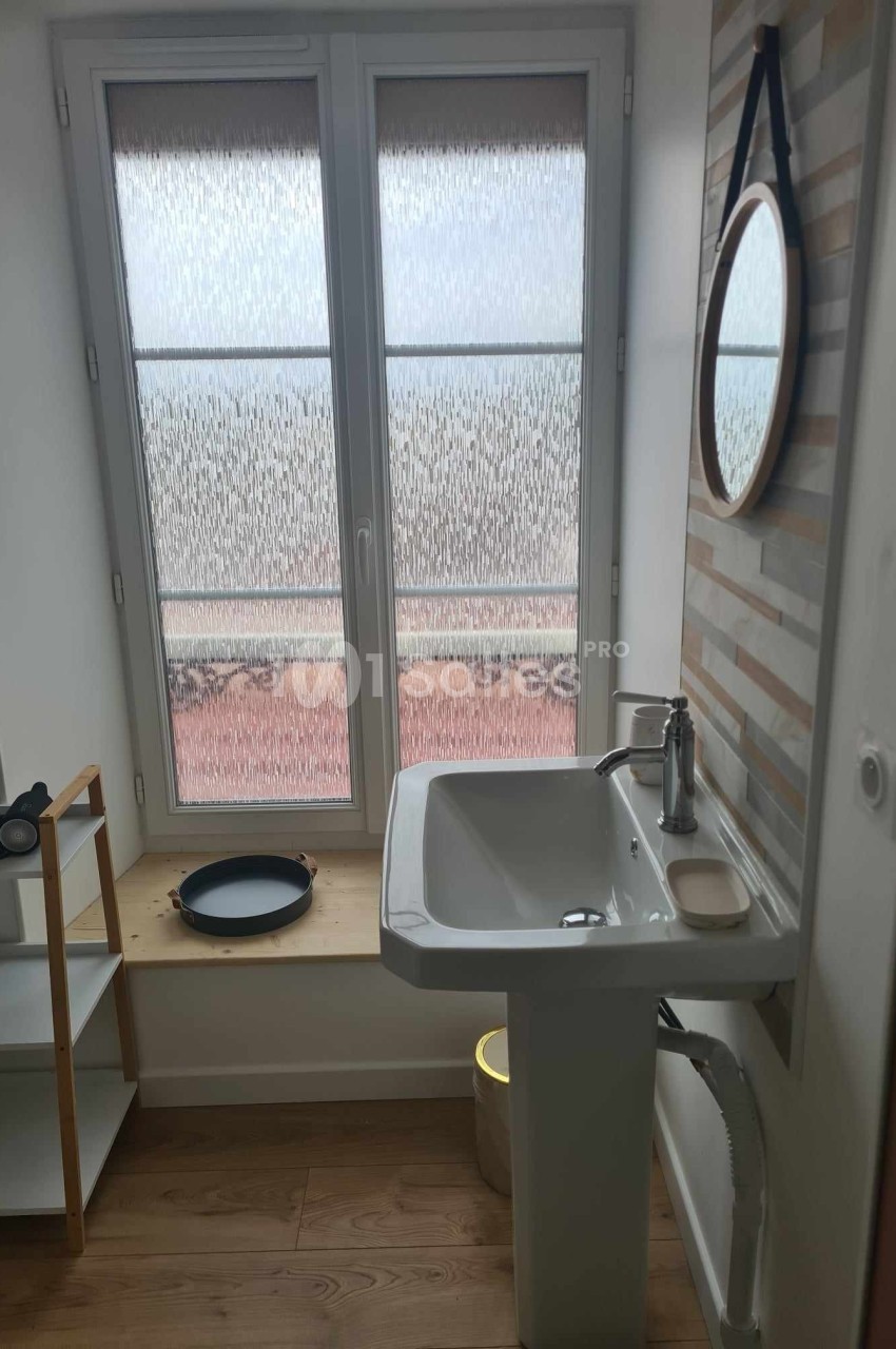 Lavabo blanc devant une fenêtre avec verre dépoli, étagère en bois à gauche et miroir rond au mur.