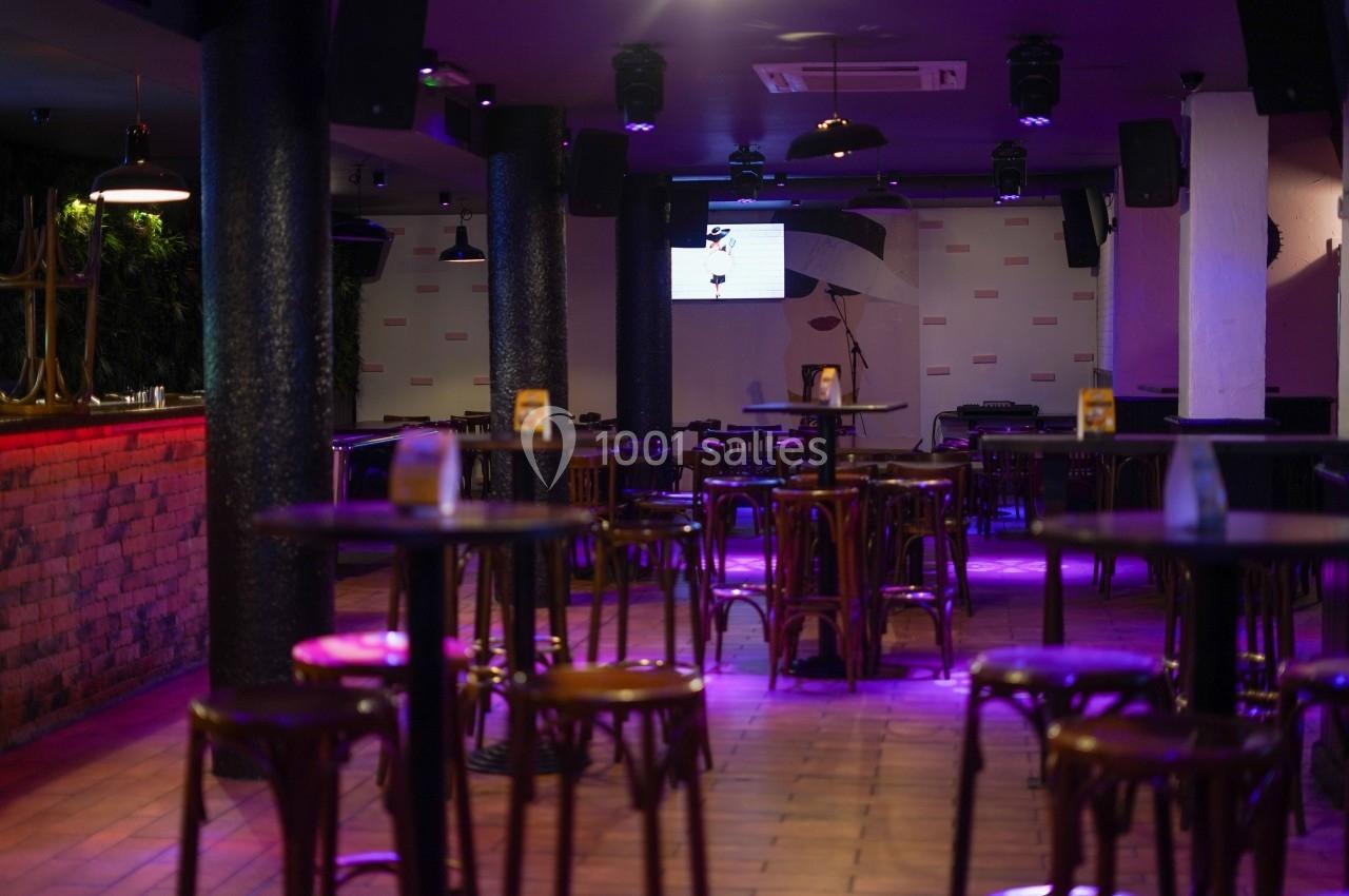 Salle de bar avec tables et chaises en bois, éclairage tamisé violet et écran au fond.