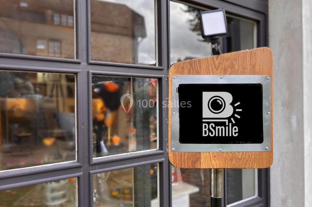 Borne photo en bois avec écran tactile devant une vitrine reflétant des bâtiments et un ciel nuageux.