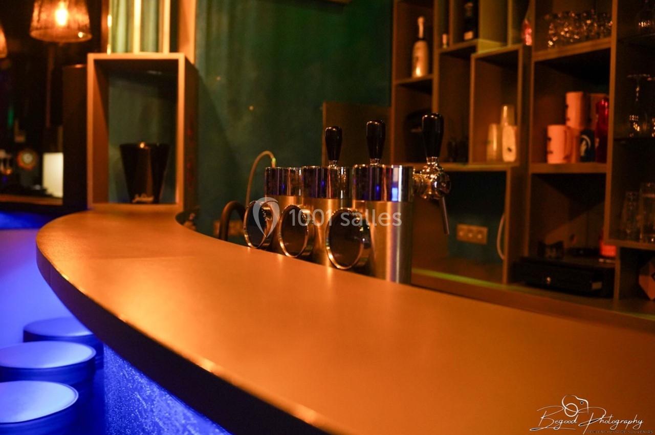 Comptoir de bar éclairé, avec tireuses à bière en métal et étagères contenant des verres et des bouteilles en arrière-plan.