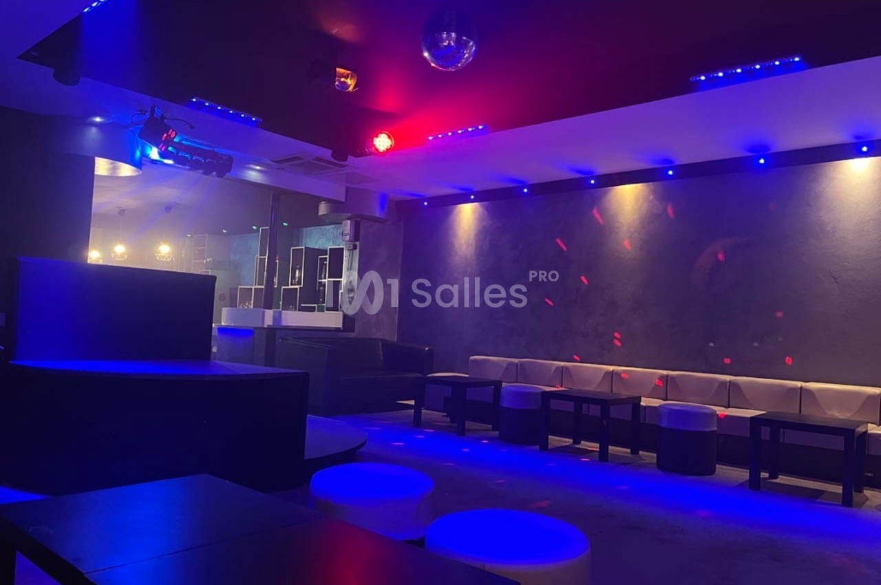 Salle de club avec éclairage tamisé bleu et rouge, banquettes, tables basses et piste de danse vide.