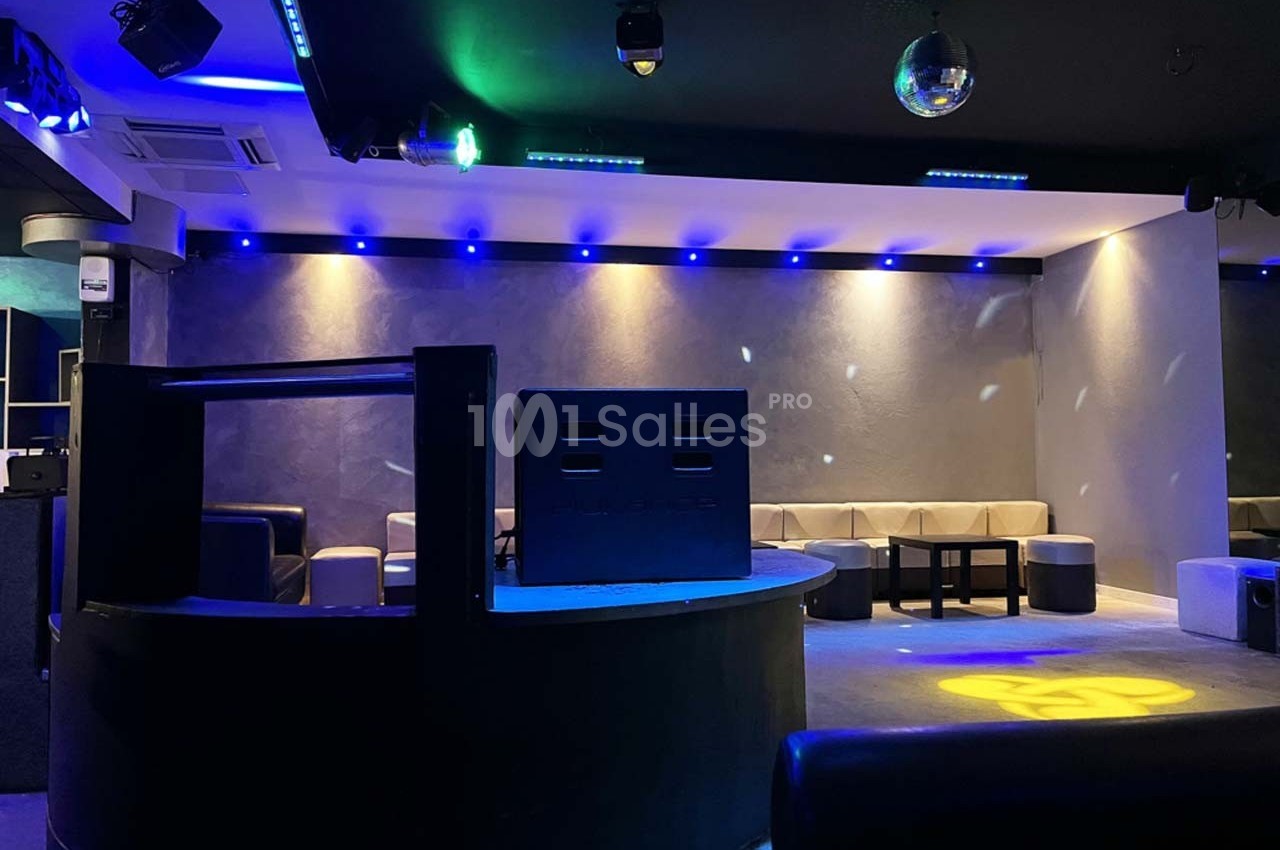 Salle de club avec éclairage bleu, banquettes grises, piste de danse et enceintes au premier plan.