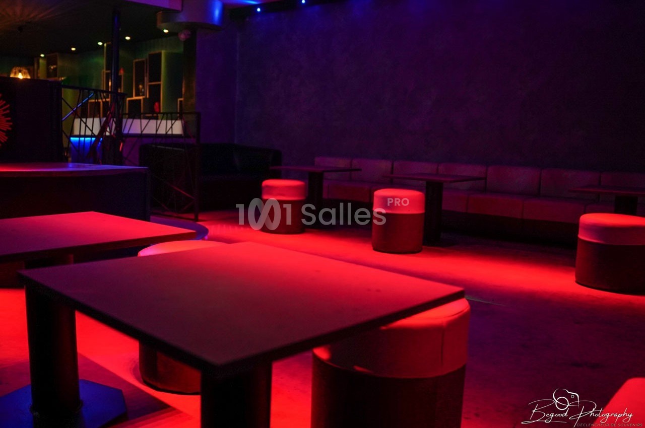 Salle de bar ou discothèque avec tables basses, tabourets et éclairage tamisé rouge et bleu.