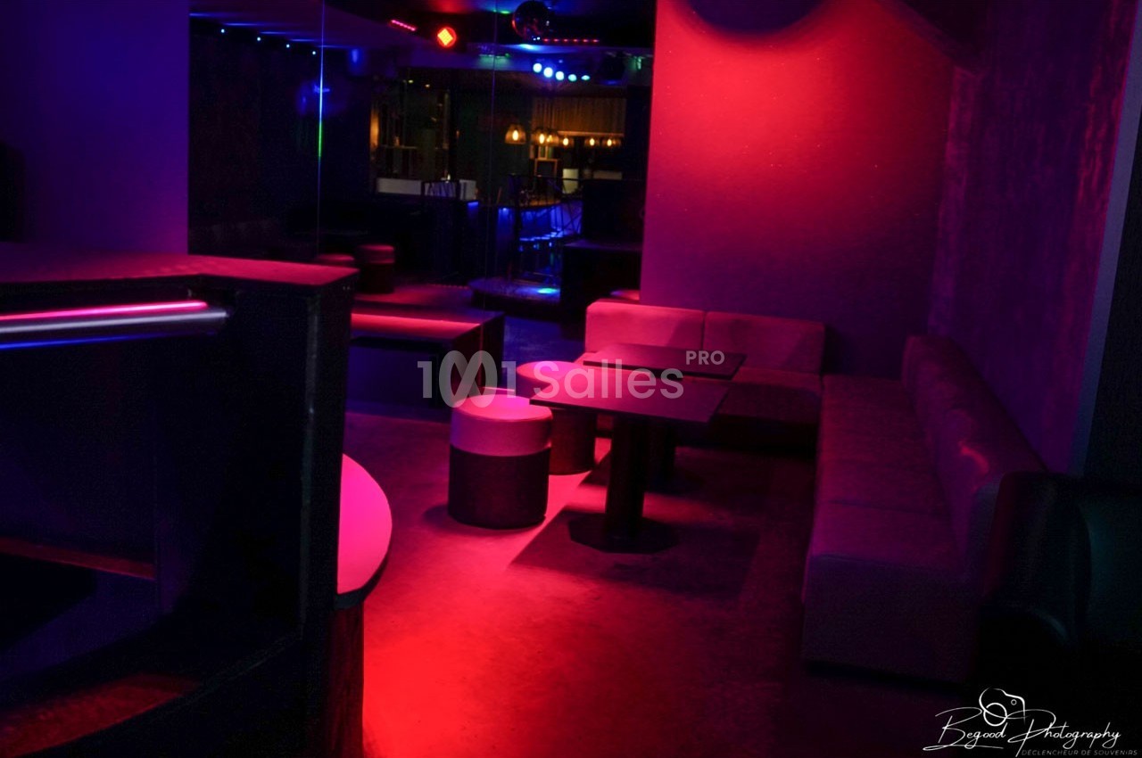 Salle de club avec éclairage tamisé rouge et violet, banquettes, tables basses et ambiance nocturne.