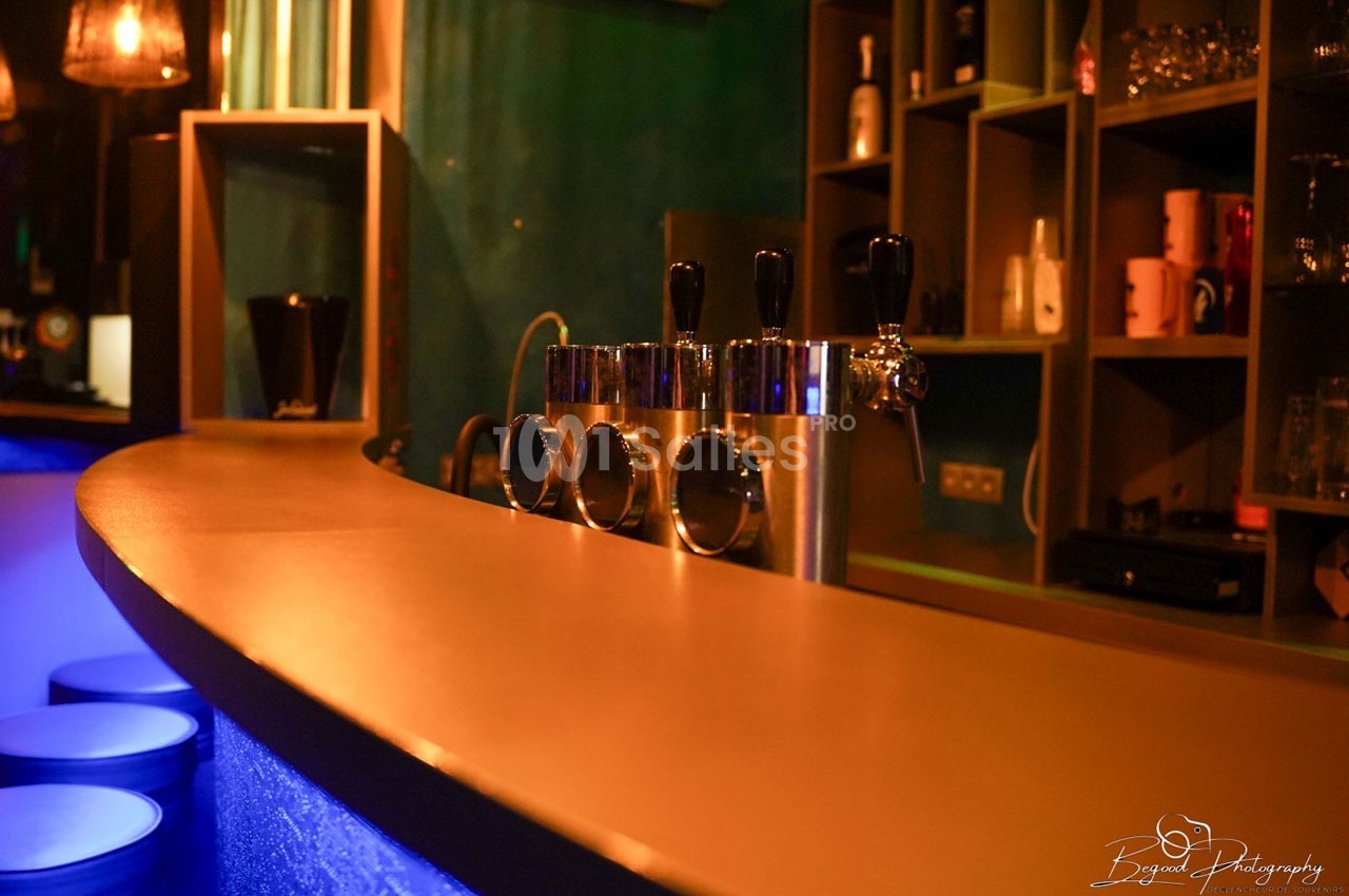 Comptoir de bar éclairé, avec tireuses à bière en métal et étagères contenant des verres et des bouteilles en arrière-plan.