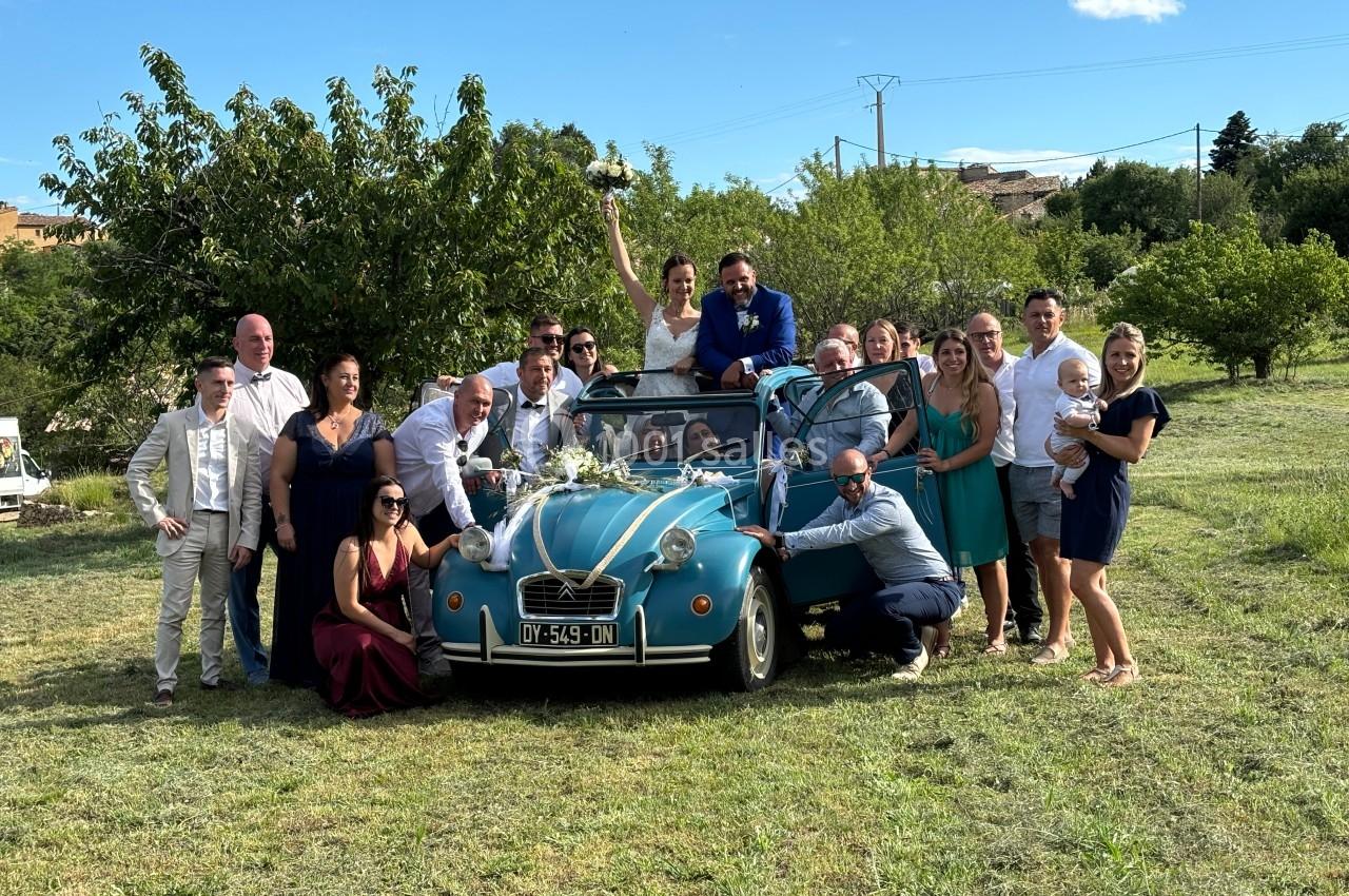 Un groupe de personnes pose autour d'une voiture ancienne décorée pour un mariage, dans un cadre champêtre.