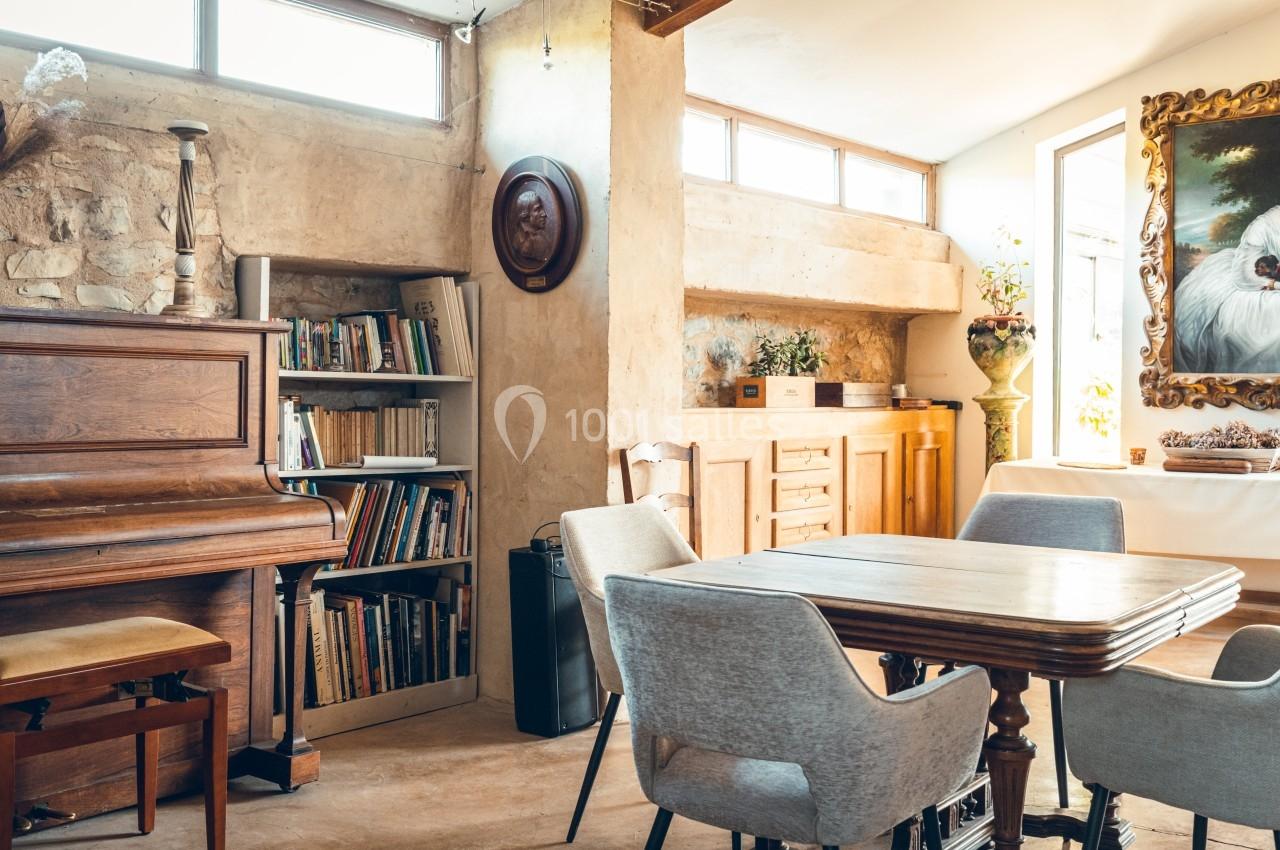 Salle à manger lumineuse avec table en bois, chaises grises, bibliothèque, piano et décoration murale.