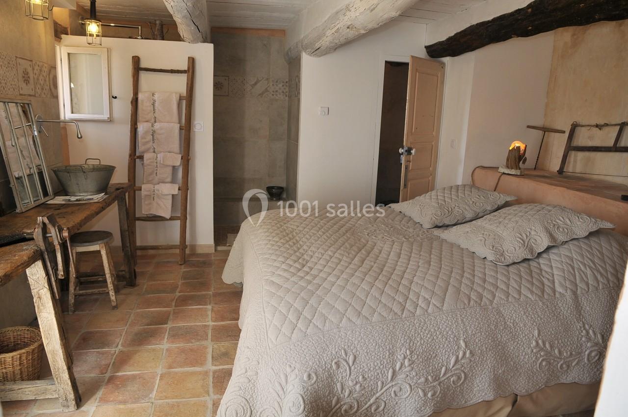 Chambre rustique avec lit double, poutres apparentes, mobilier en bois et salle de bain attenante en arrière-plan.