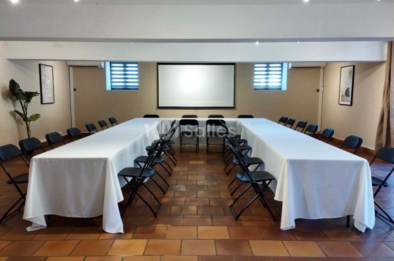 Salle de réunion avec tables en U couvertes de nappes blanches, chaises noires et écran de projection en fond.