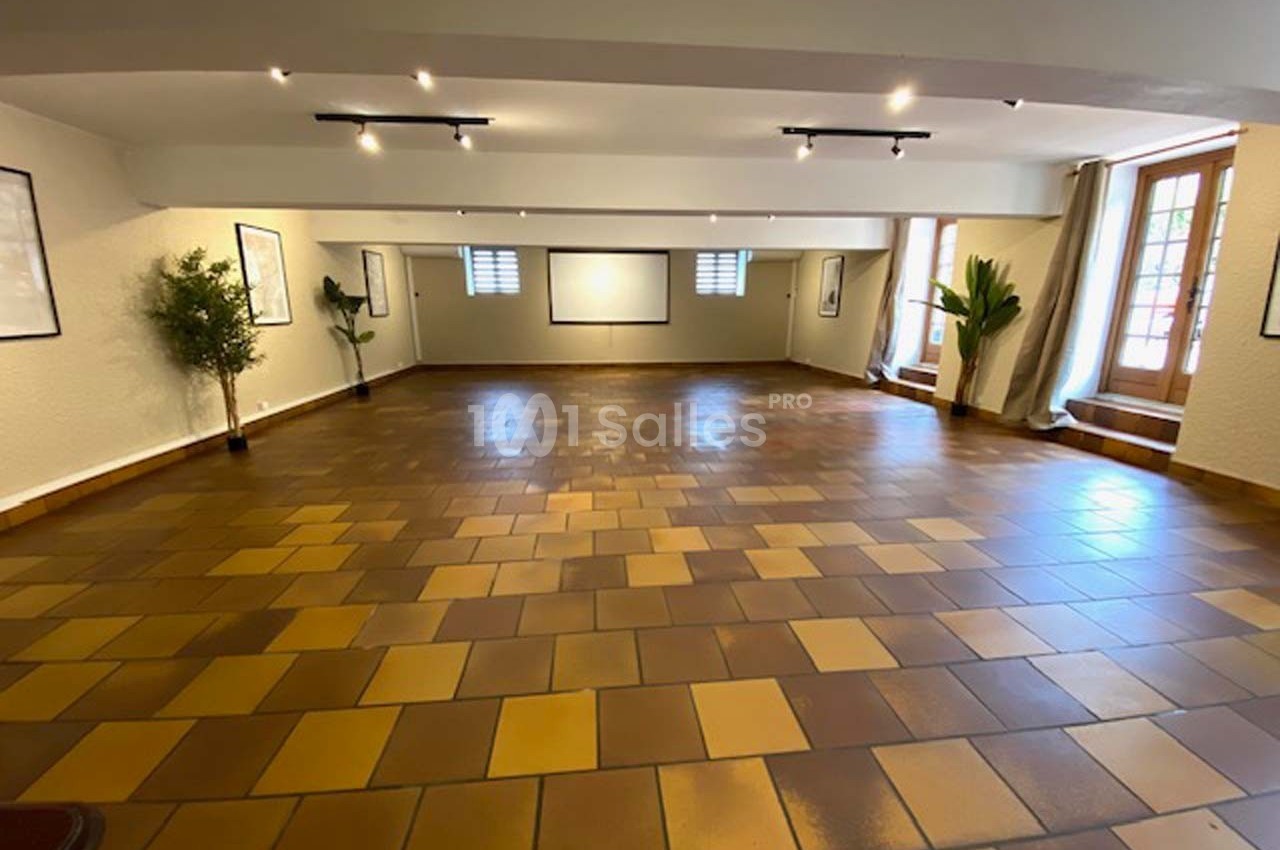 Salle vide avec sol en carrelage marron, murs clairs, grandes fenêtres, plantes et éclairage au plafond.