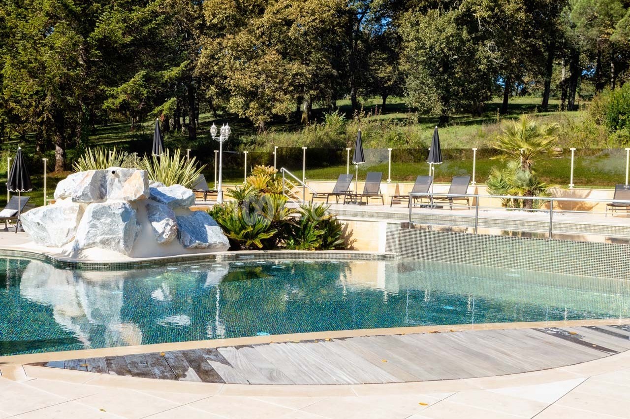 Piscine extérieure entourée de végétation, transats et parasols noirs, avec des rochers décoratifs au premier plan.