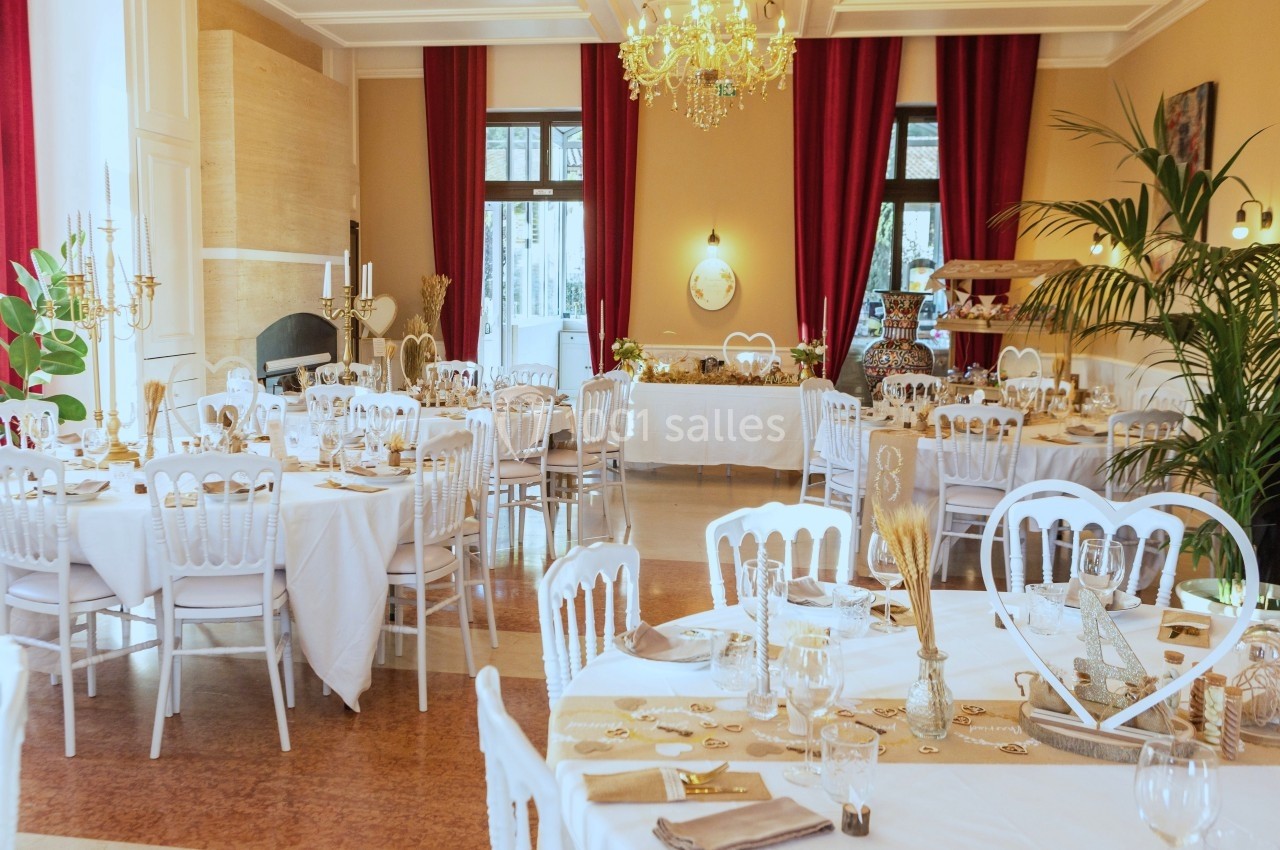 Salle de réception élégante avec tables dressées, chaises blanches, décorations dorées et rideaux rouges.