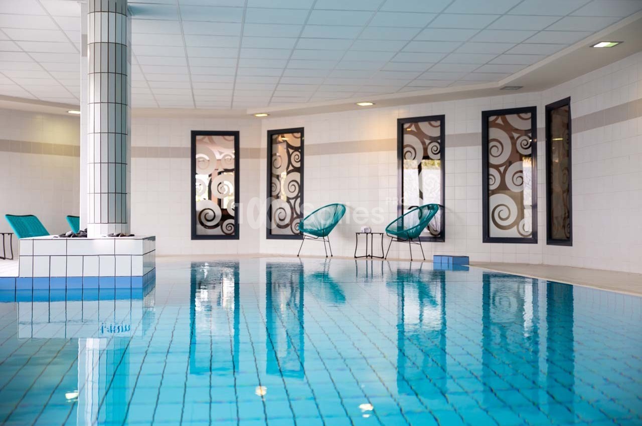 Piscine intérieure avec carrelage bleu, chaises modernes et grandes fenêtres ornées de motifs décoratifs.