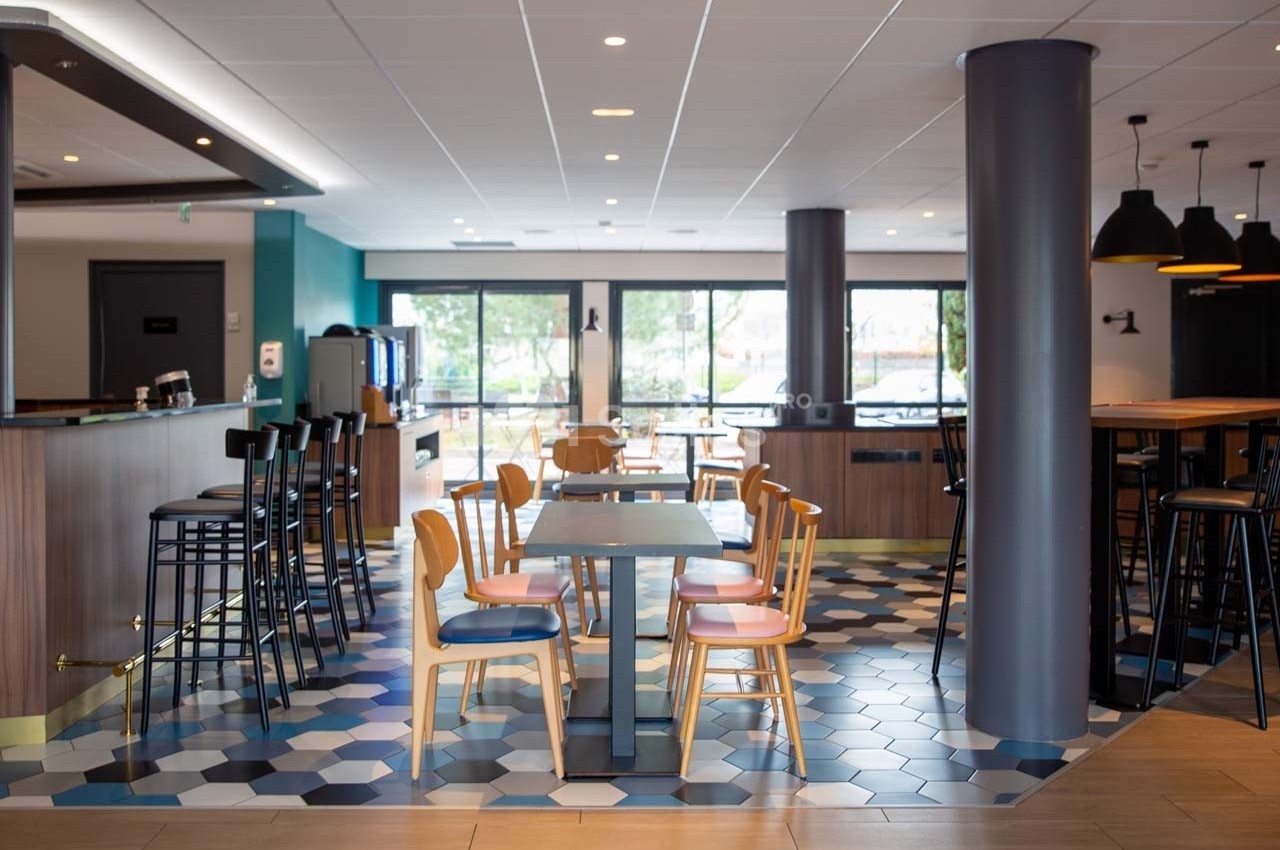 Salle de restaurant moderne avec tables, chaises en bois et carrelage hexagonal, éclairée par de grandes baies vitrées.