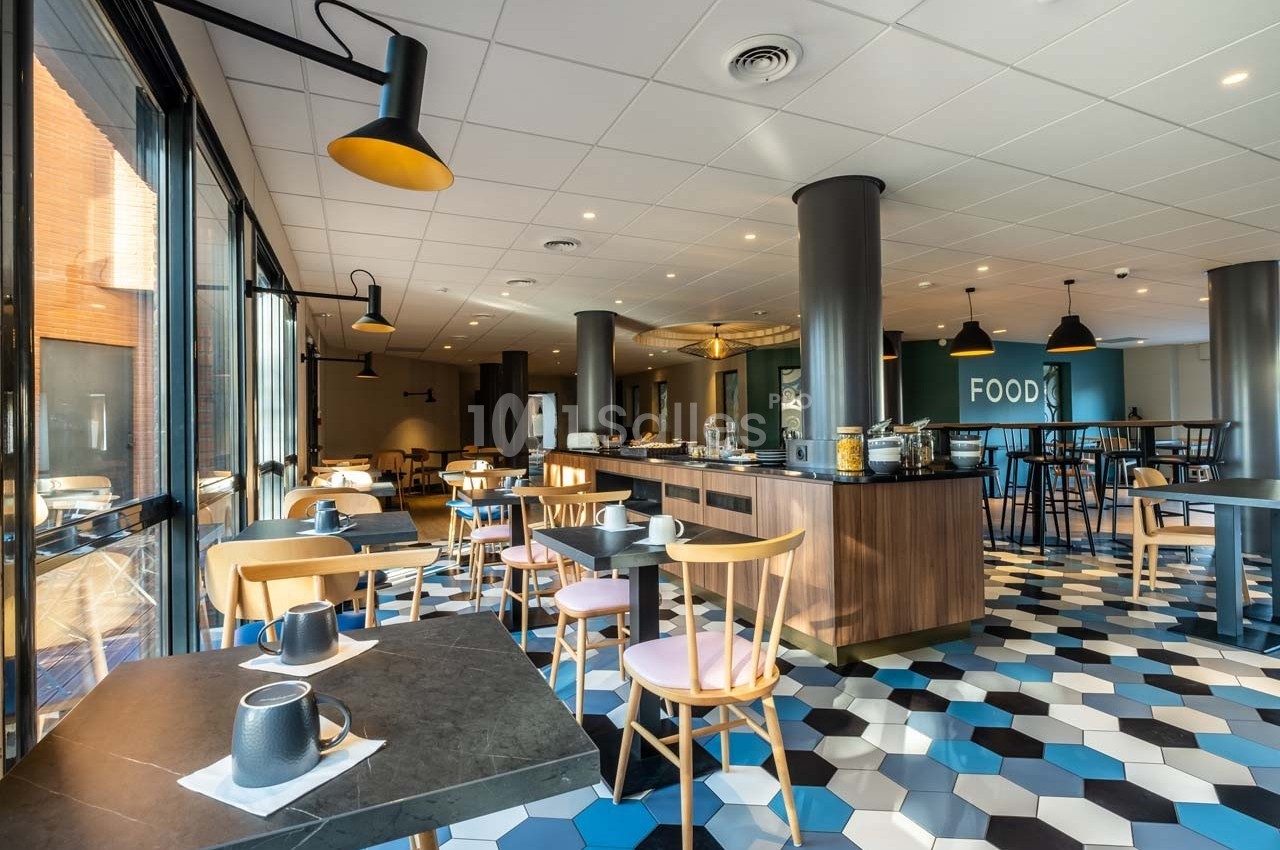 Salle de restaurant lumineuse avec tables, chaises en bois, sol à motifs géométriques et buffet central.