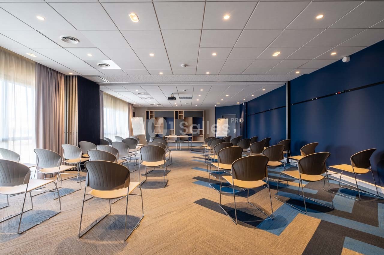 Salle de conférence moderne avec des chaises disposées en rangées, éclairée par des fenêtres et des lumières au plafond.
