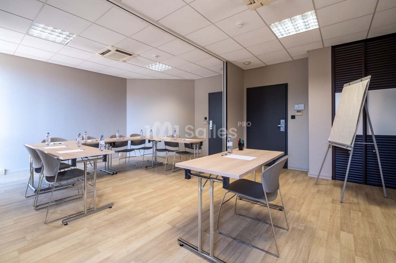 Salle de réunion lumineuse avec tables, chaises, tableau blanc et bouteilles d'eau sur un sol en parquet clair.