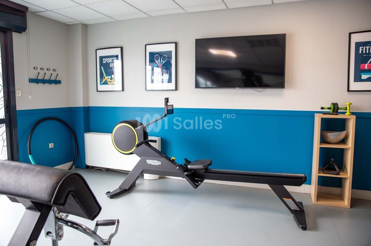 Salle de sport avec rameur, écran mural, affiches encadrées et équipement de fitness rangé sur des étagères.