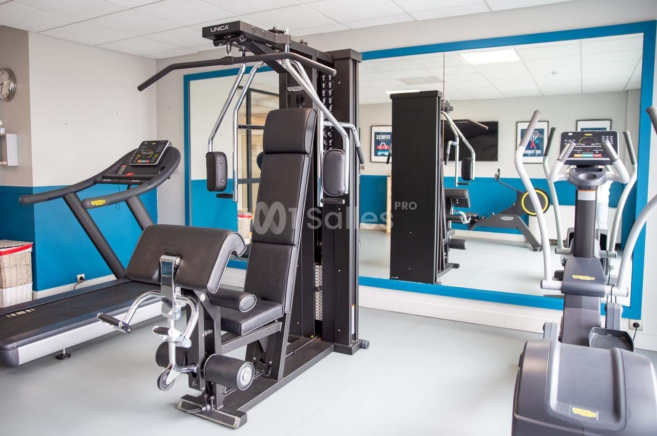 Salle de sport équipée de machines de musculation, tapis de course et vélo elliptique, avec miroir mural.