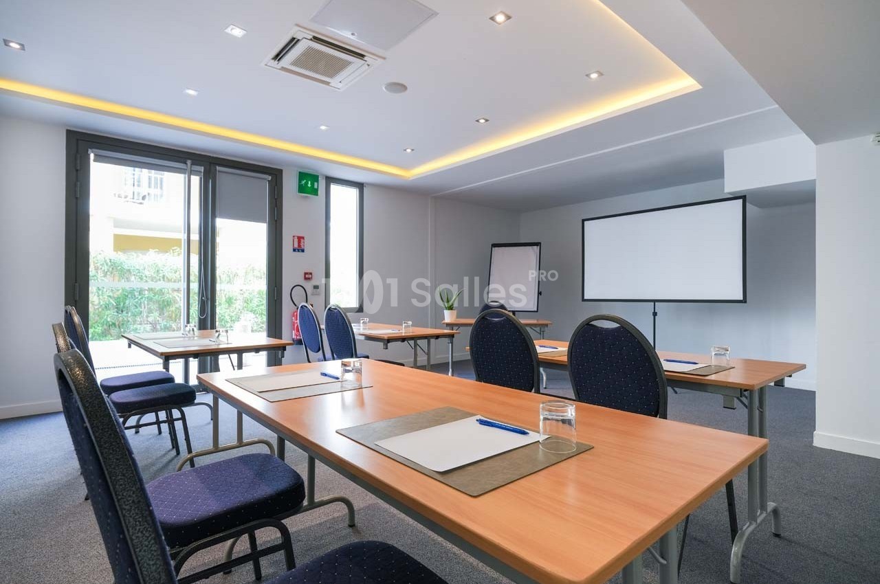 Salle de réunion lumineuse avec tables en bois, chaises, tableau blanc et écran de projection.
