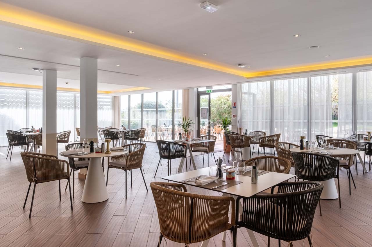 Salle de restaurant lumineuse avec des tables dressées, des chaises en rotin et de grandes baies vitrées.