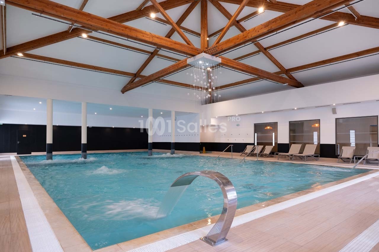 Piscine intérieure avec jets d'eau, transats alignés sur le côté et plafond en poutres apparentes.