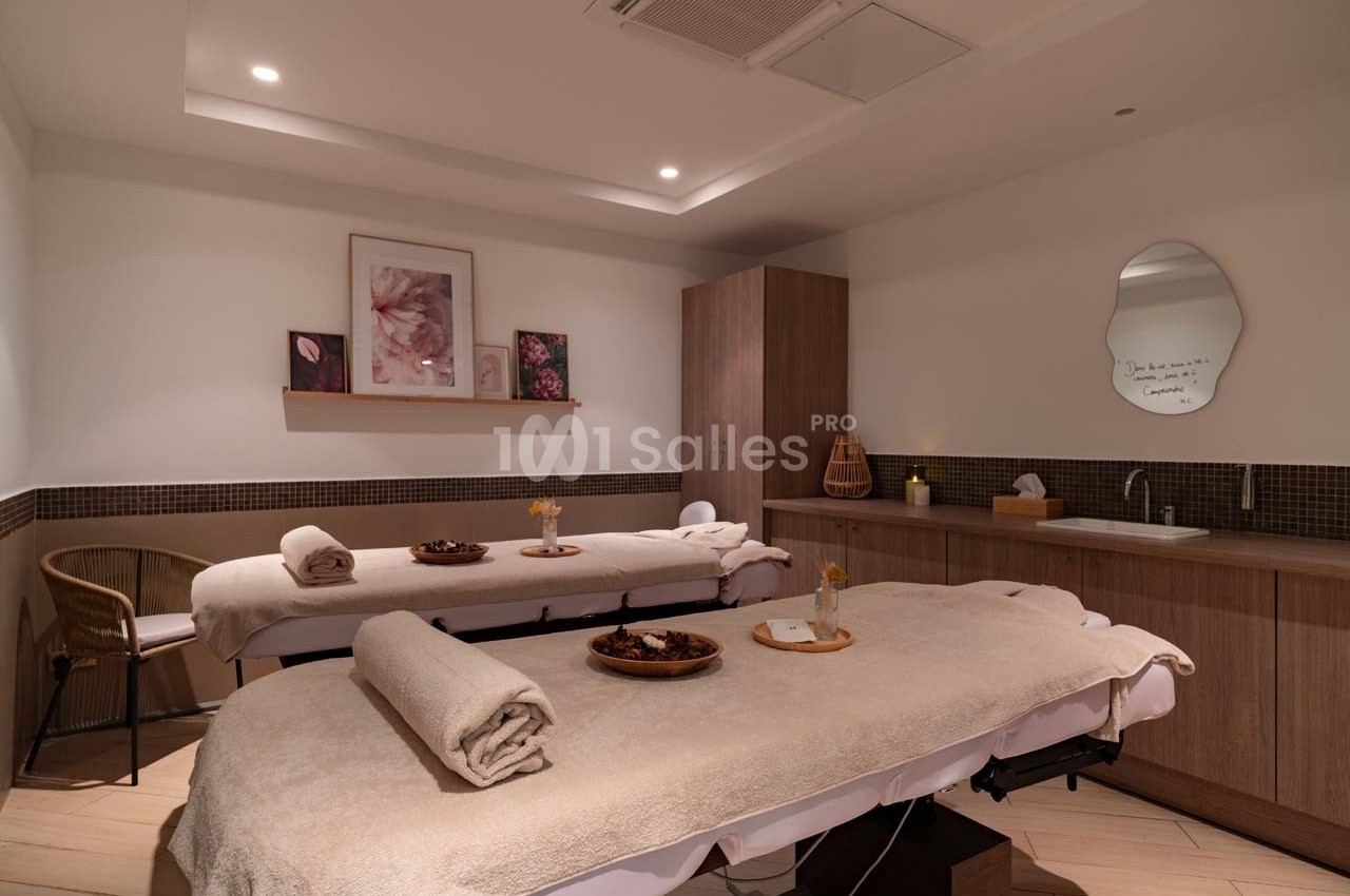 Deux tables de massage avec serviettes et décor apaisant dans une salle lumineuse et aménagée pour des soins.