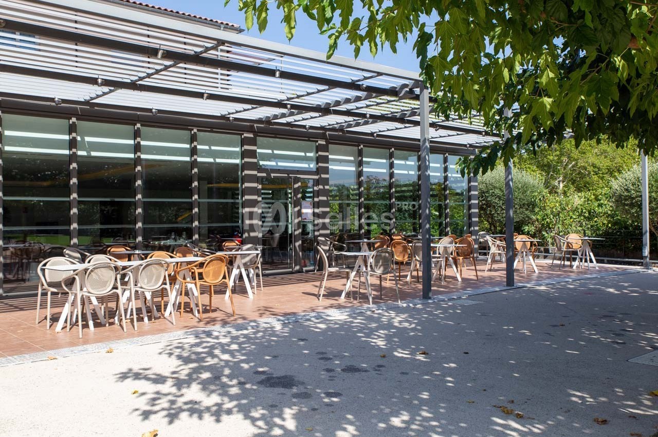 Terrasse extérieure avec des tables et chaises vides, ombragée par des arbres et une pergola, près d'un bâtiment vitré.