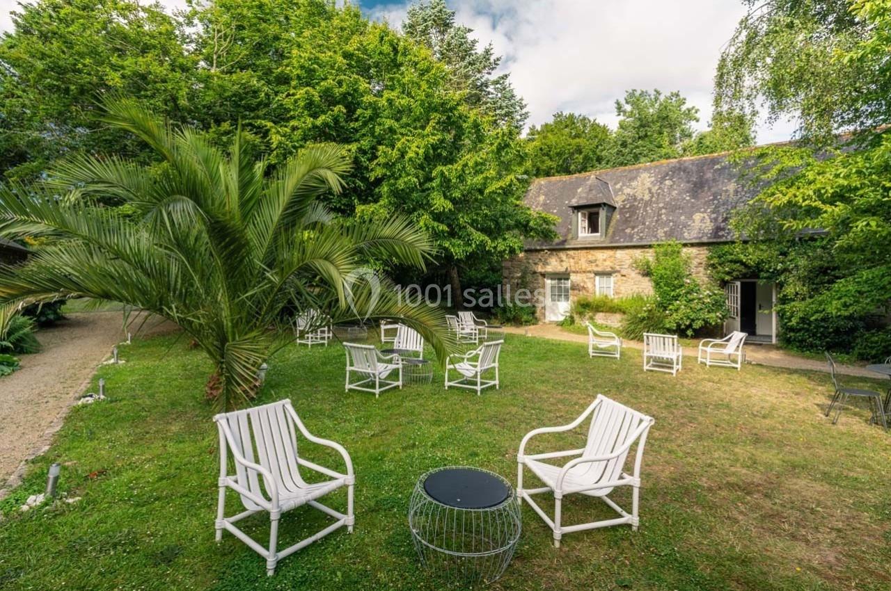 Jardin verdoyant avec fauteuils blancs disposés autour de petites tables, devant une maison en pierre sous un ciel…