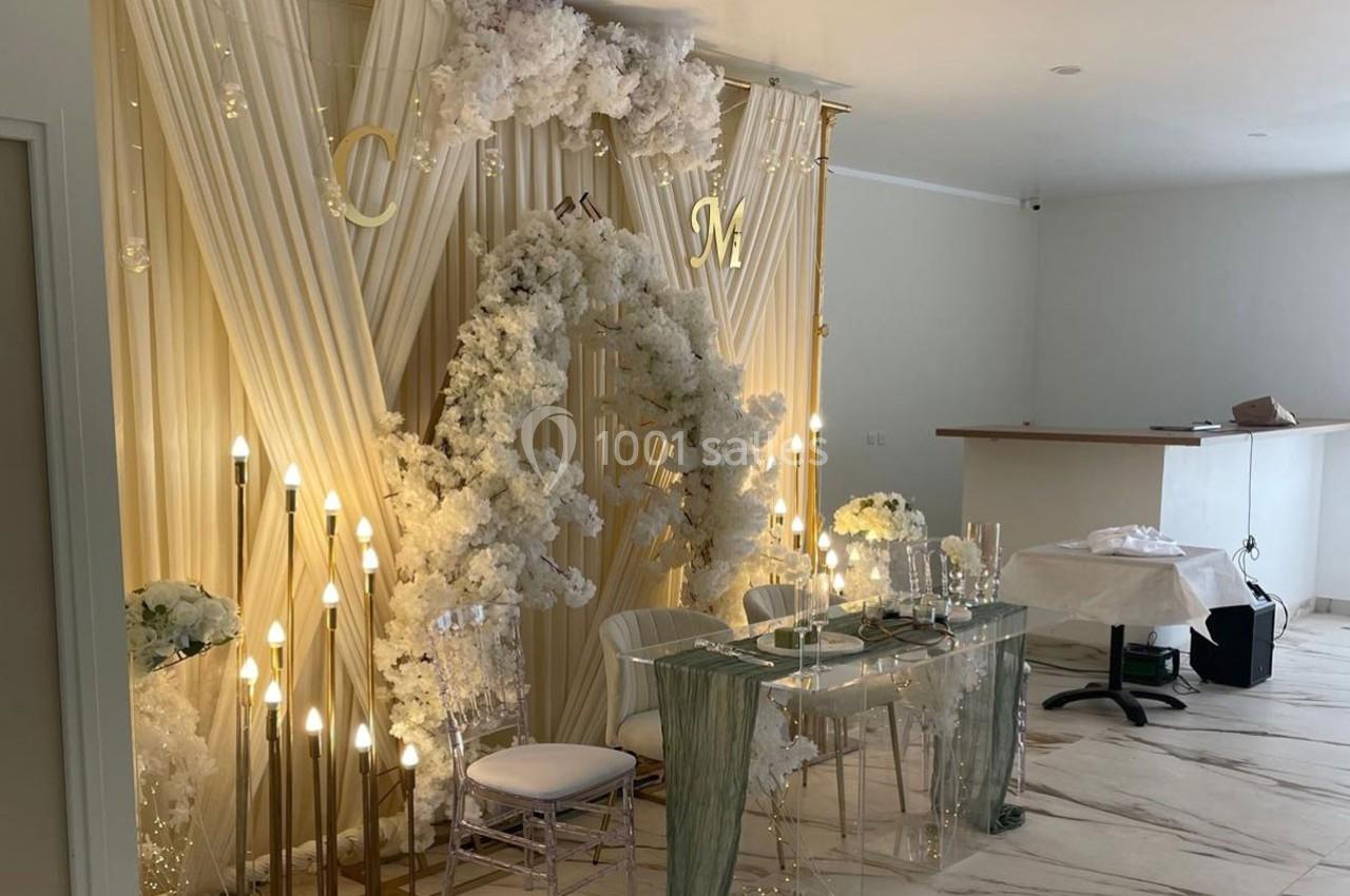 Décoration de cérémonie avec rideaux blancs, arche florale blanche, éclairage doré et table transparente dressée.