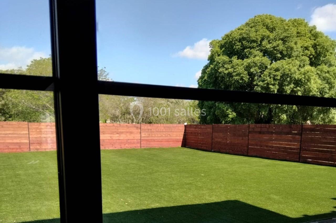 Vue depuis une fenêtre sur un jardin avec pelouse artificielle, clôture en bois et arbres en arrière-plan.