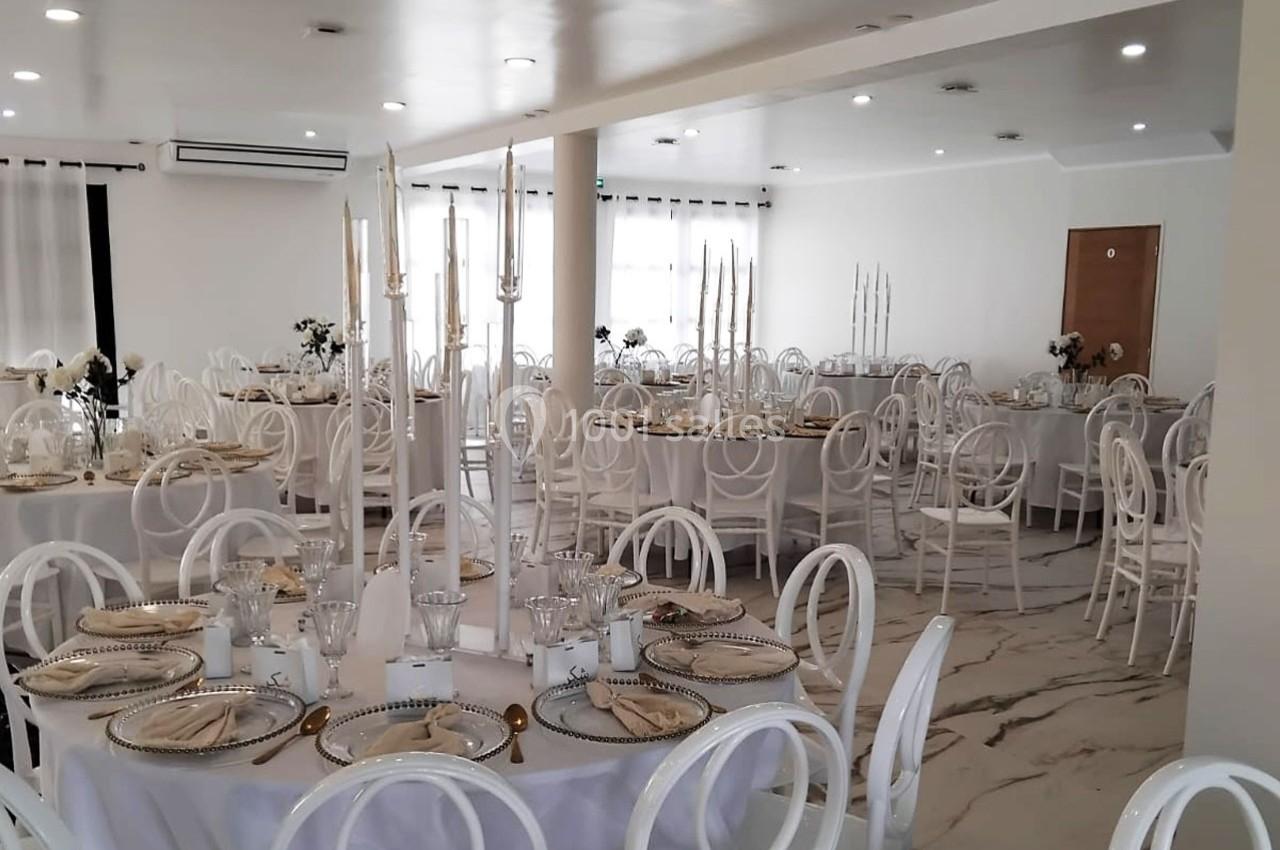 Salle de réception lumineuse avec tables rondes dressées, chaises blanches et chandeliers élégants.