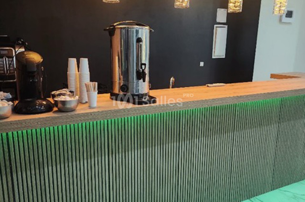 Comptoir en bois avec machine à café, distributeur d'eau chaude, gobelets et éclairage suspendu dans un espace moderne.