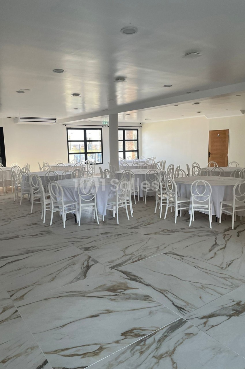 Salle lumineuse avec tables rondes dressées de nappes blanches et chaises blanches sur un sol en marbre clair.