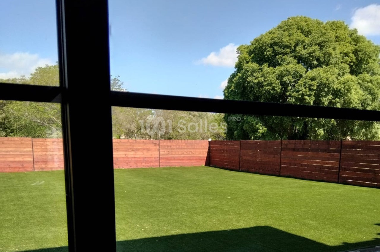 Vue depuis une fenêtre sur un jardin avec pelouse artificielle, clôture en bois et arbres en arrière-plan.