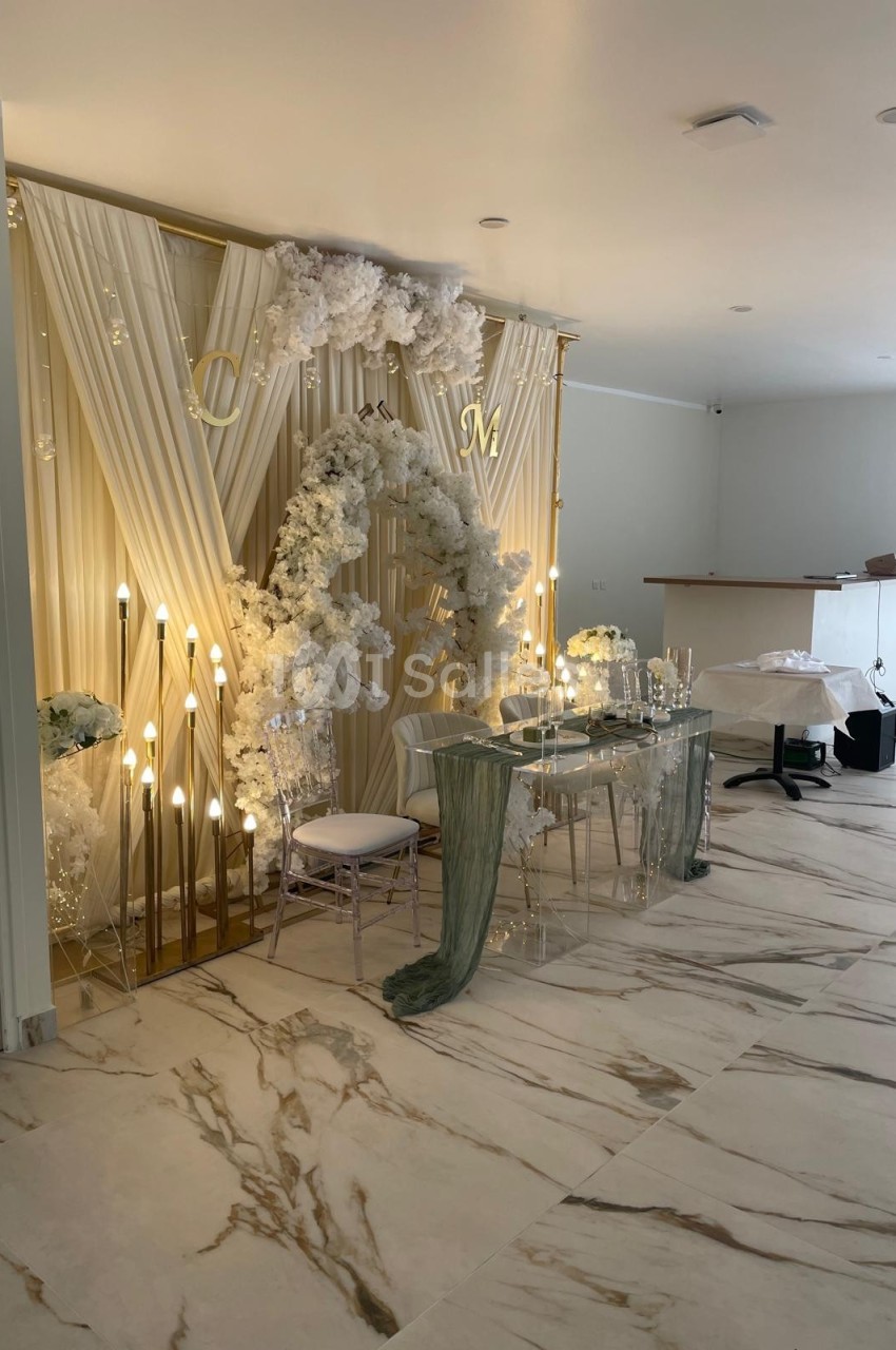 Décoration de mariage élégante avec arche florale blanche, rideaux drapés, guirlandes lumineuses et table transparente.