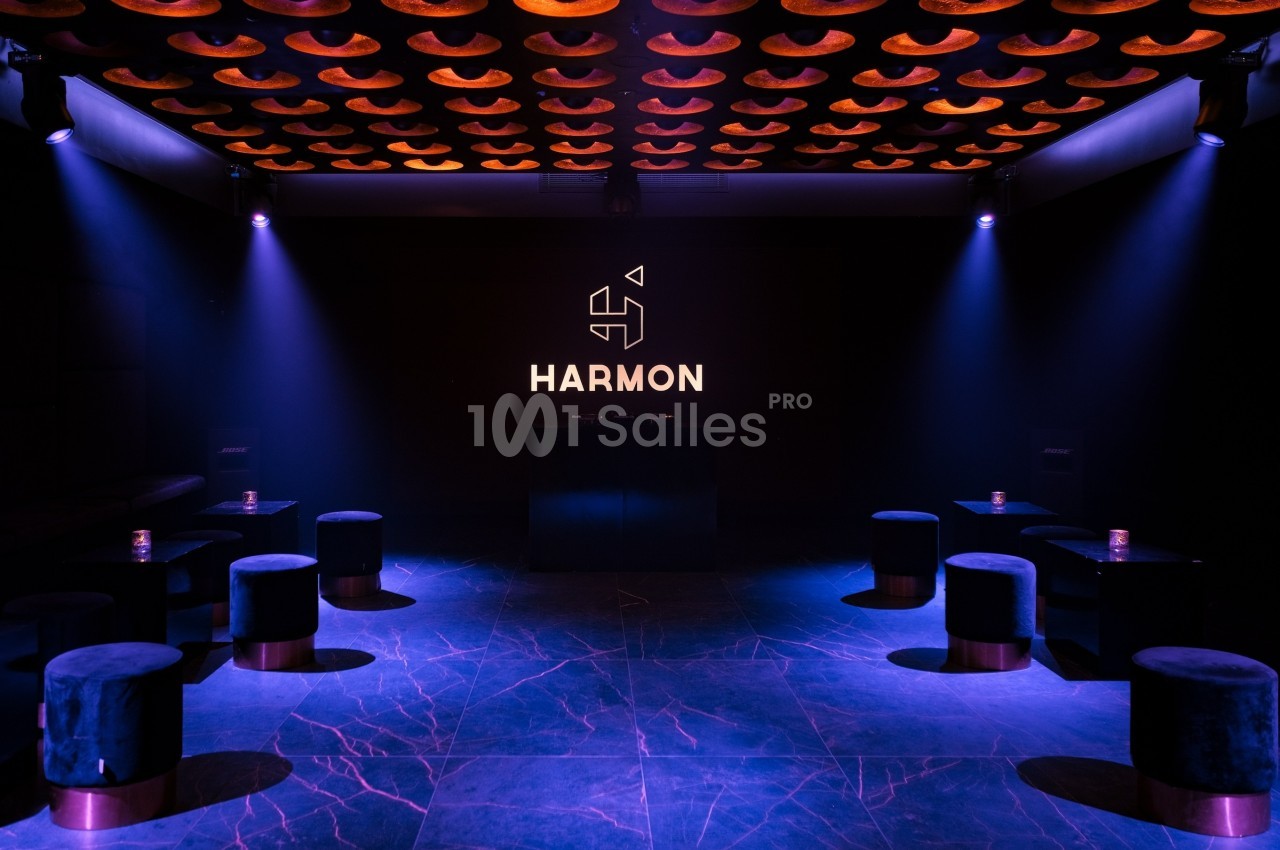 Salle sombre avec éclairage violet, logo ’Harmon’ illuminé sur un mur noir, tabourets disposés autour de petites tables.