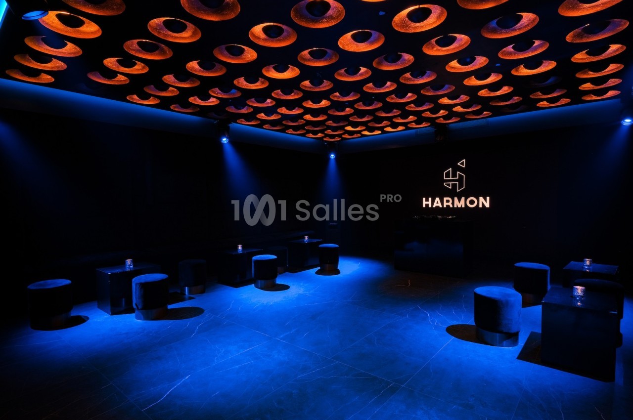 Salle sombre avec un éclairage bleu, plafond orné de cercles lumineux orange et logo ’Harmon’ illuminé sur un mur.