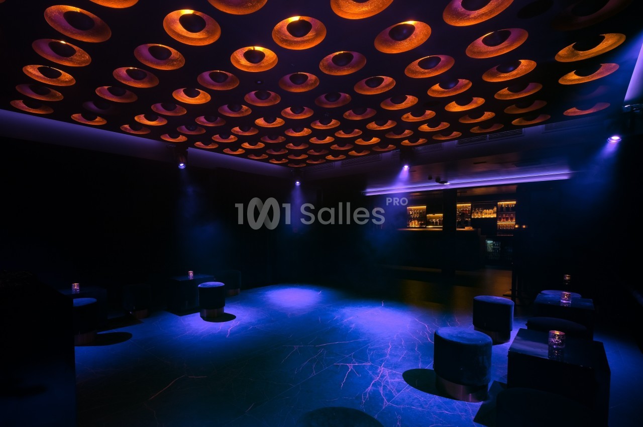 Salle sombre avec un plafond décoratif circulaire illuminé, des tabourets noirs et un bar éclairé en arrière-plan.
