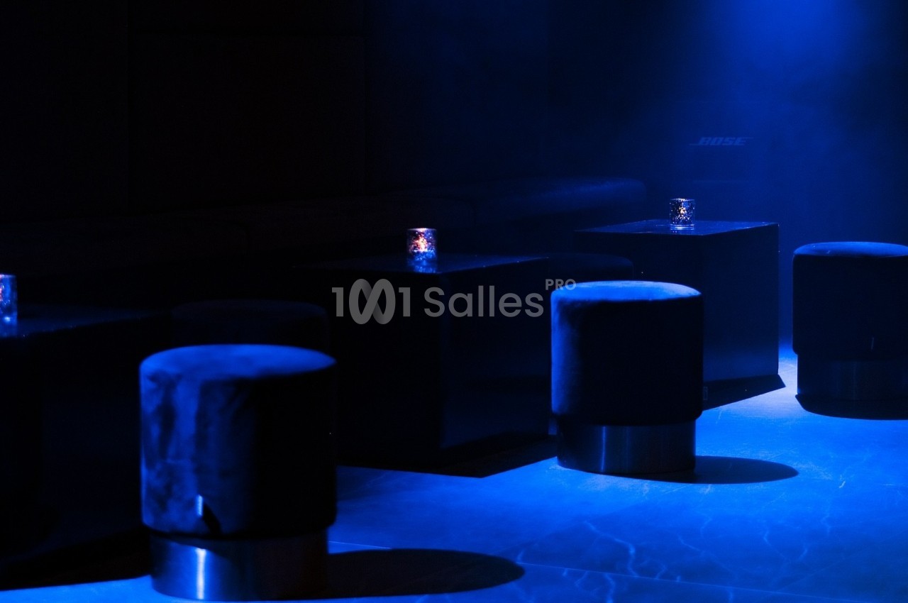 Tabourets noirs et tables basses éclairés par une lumière bleue tamisée dans un espace sombre.