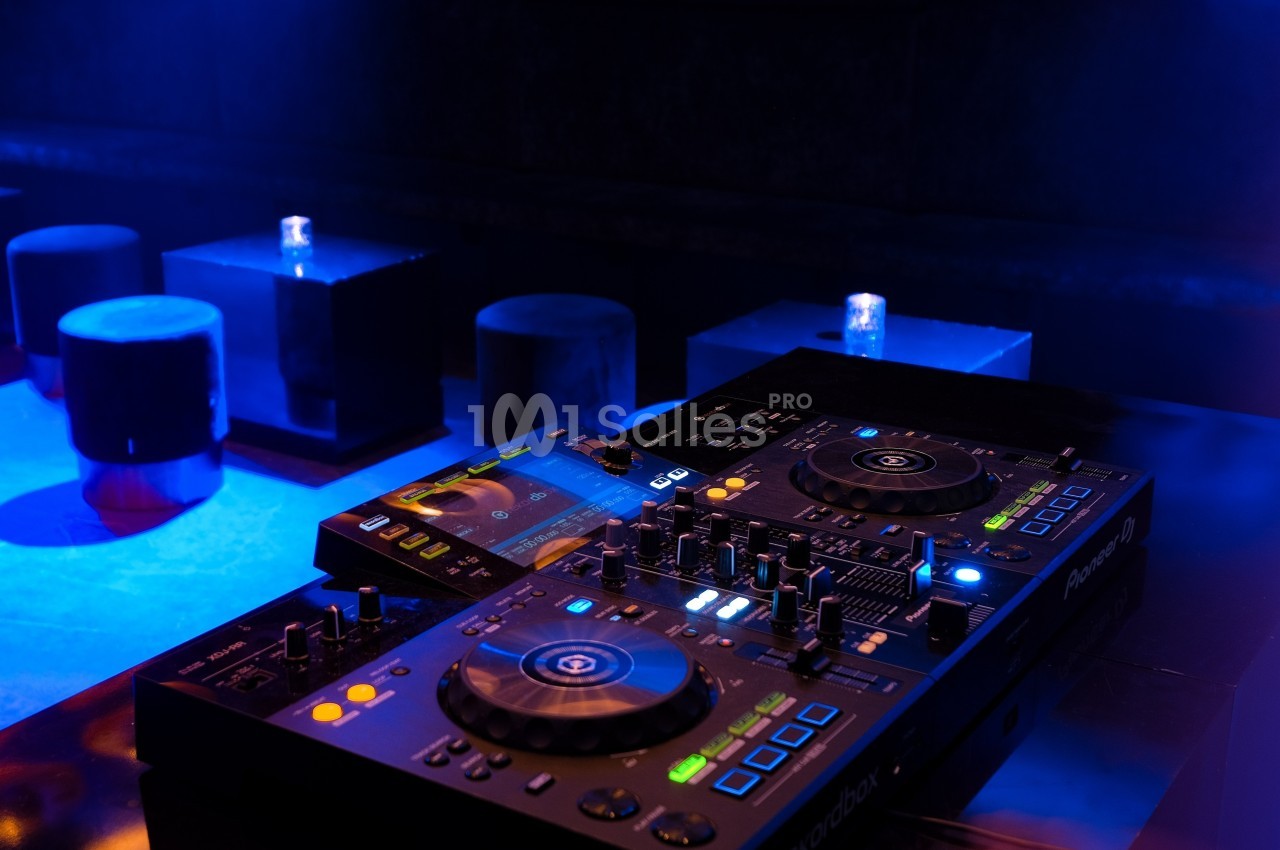 Table de mixage DJ éclairée dans une ambiance sombre, avec des tabourets et tables en arrière-plan.