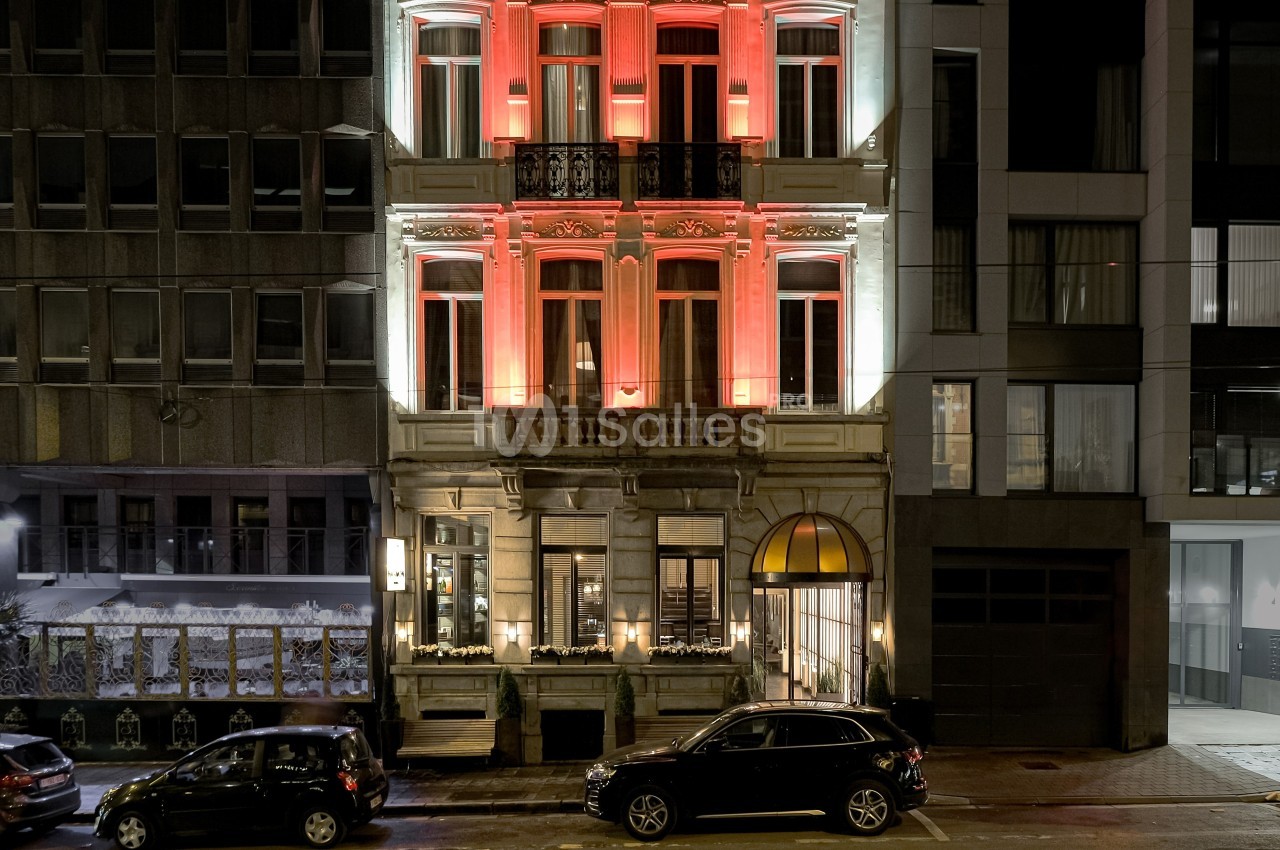 Façade d'un immeuble ancien éclairé en rouge et blanc, entouré de bâtiments modernes, avec des voitures stationnées devant.