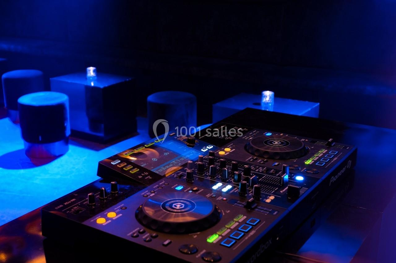 Table de mixage DJ éclairée dans une ambiance de club avec des tabourets et des tables en arrière-plan.