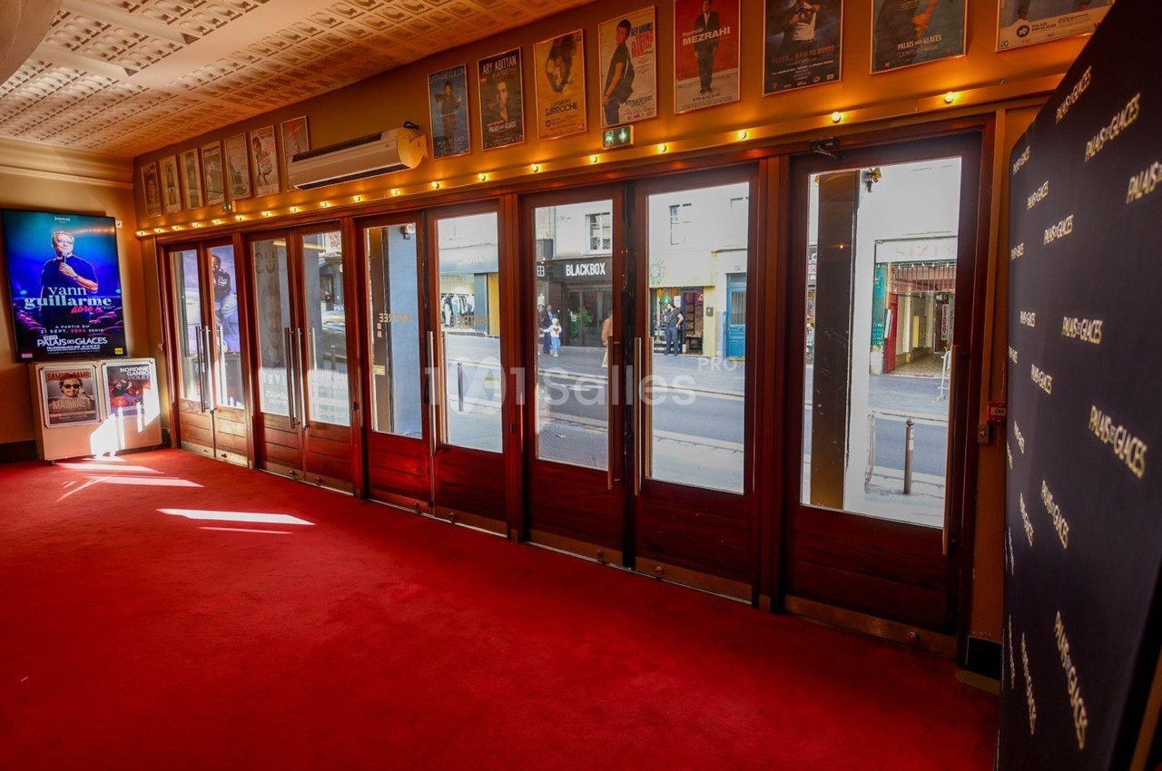 Entrée vitrée d'un théâtre avec tapis rouge, affiches de spectacles au mur et vue sur la rue à travers les portes.