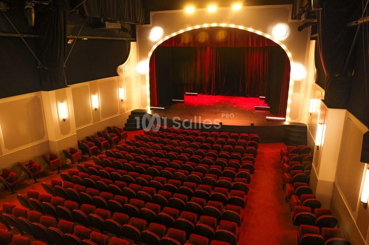 Salle de théâtre vide avec des rangées de sièges rouges, une scène éclairée et un rideau rouge fermé.