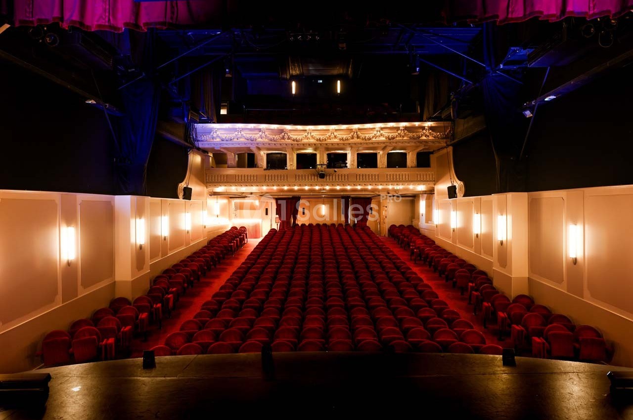 Salle de théâtre vide avec rangées de sièges rouges, vue depuis la scène, éclairage tamisé et balcon illuminé.