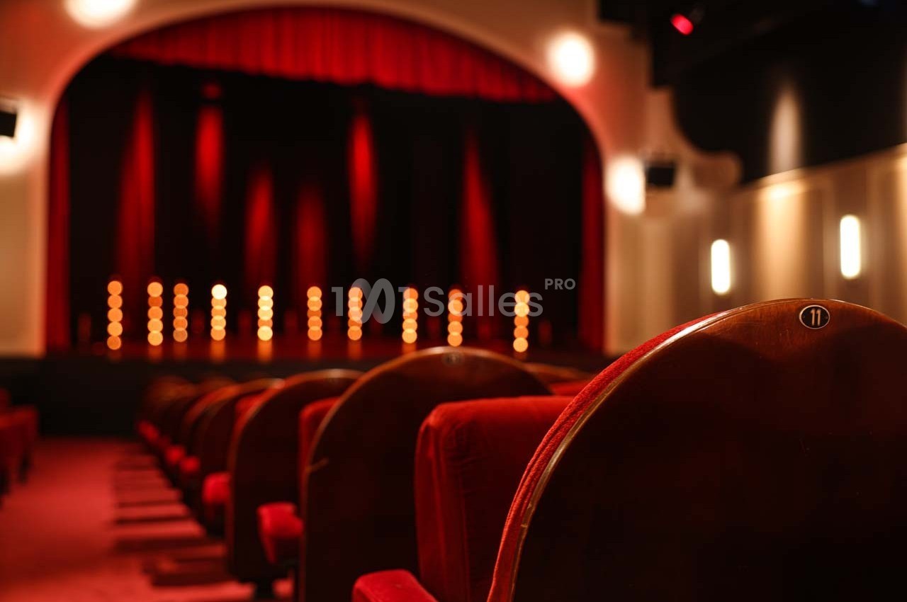 Salle de théâtre avec des sièges rouges au premier plan et une scène éclairée par des lumières en arrière-plan.