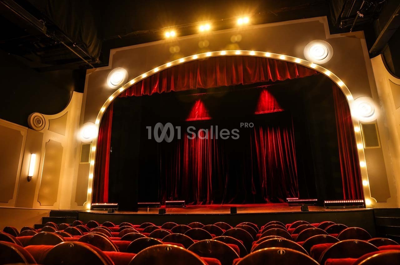 Salle de théâtre avec rideaux rouges fermés, éclairée par des projecteurs et entourée de sièges en velours rouge.