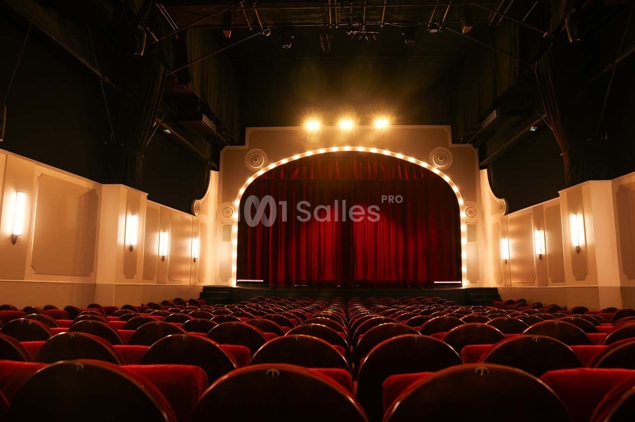 Salle de théâtre vide avec rangées de sièges rouges et une scène éclairée par des projecteurs.