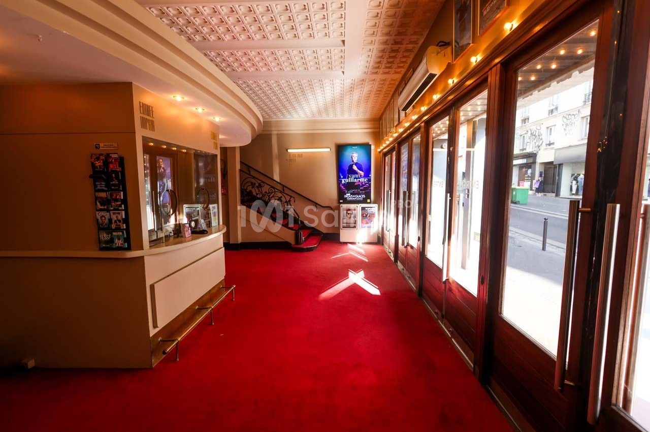 Hall d'entrée d'un théâtre avec tapis rouge, guichet à gauche, escalier central et portes vitrées donnant sur la rue.